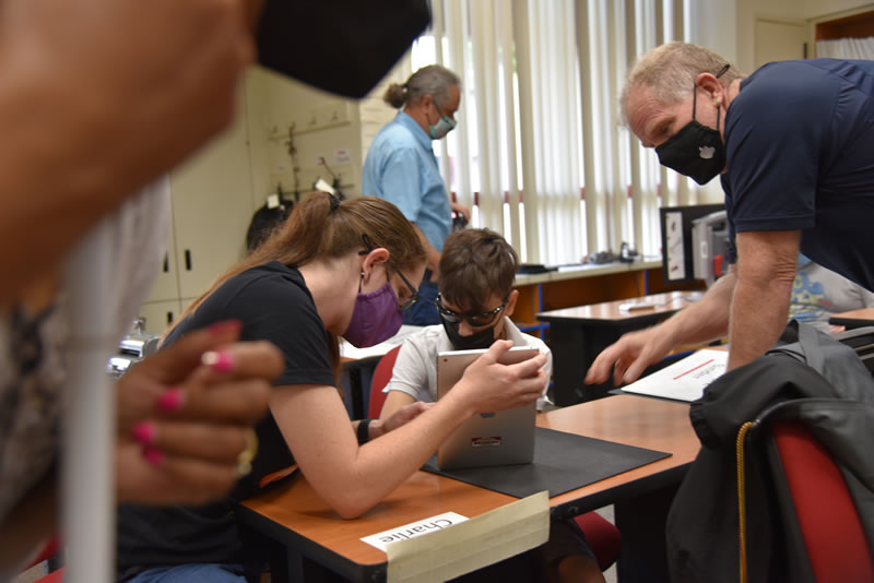 A student sitting at a desk in a classroom using an iPad with the assistance of a two adults.