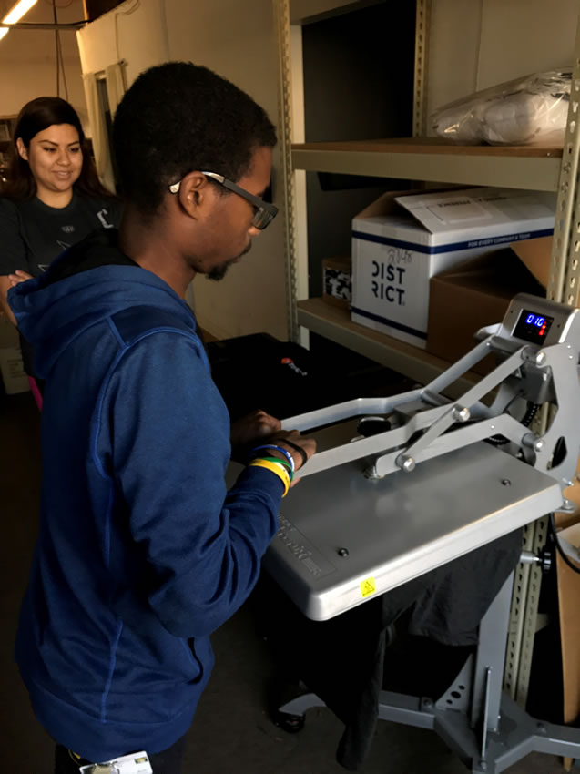 A student is pressing a design into a t shirt using machinery under supervision of an adult.