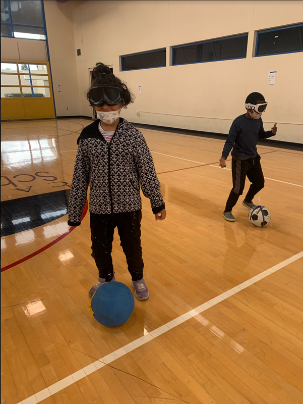 Two students are wearing blindfolds, each having a ball at their feet and practicing dribbling soccer balls. 