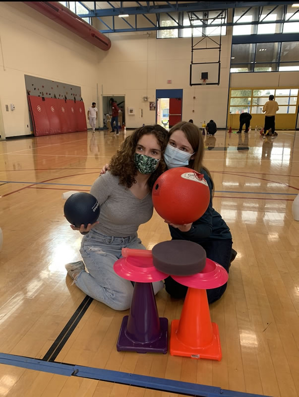 2 students are kneeling on the ground, the student on the left is holding a small ball, the student on the right is holding a beep kickball. In front of them are some cones with plates and a racket balanced on top.