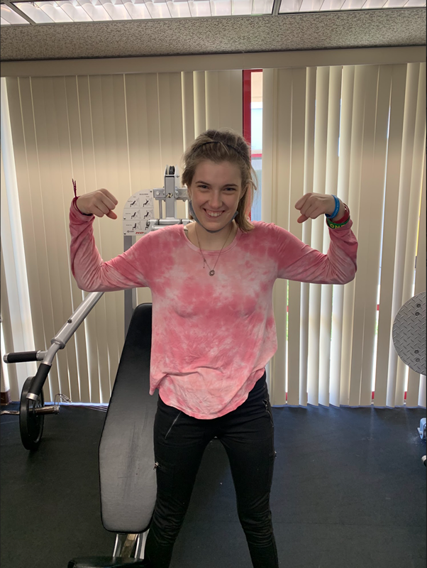 A student wearing a pink tie dye shirt is standing in front of a shoulder press machine. She has her arms up, hands in fists while smiling to show off her muscles.
