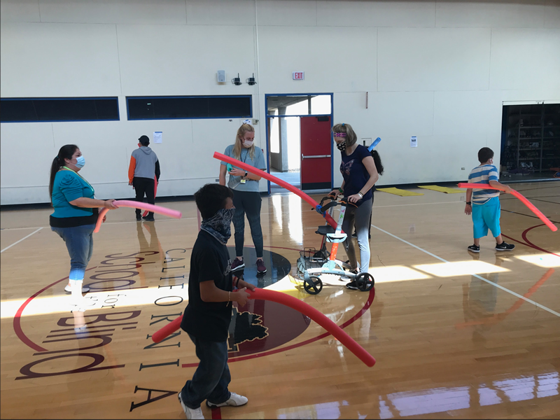 A group of 4 students and 2 teachers are moving around the gym with pool noodles.
