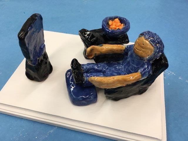 Ceramic person sitting in a chair, feet resting on a foot stool, and facing a ceramic TV.