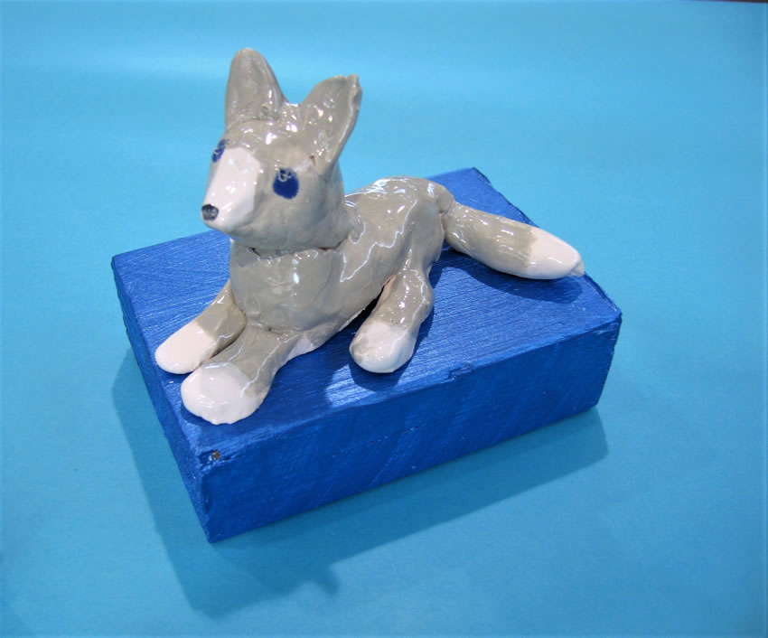 Ceramic sculpture of a wolf, lying down on a rectangle shaped, blue, wooden base.