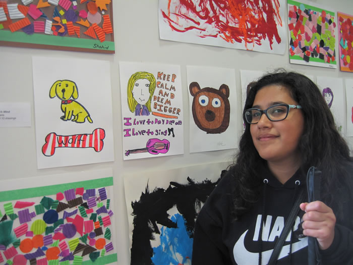 Student standing next to her drawings of a dog with a bone, a girl with guitar, and a bears face.