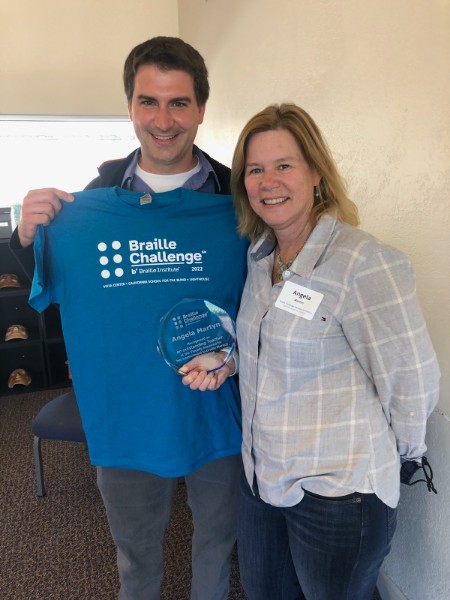 Adrian holding a Braille Challenge t-shirt standing next to Angela holding a nonagon plaque.