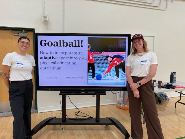 Molly and Taylor stand smiling on either side of a large screen in a gymnasium. The screen displays the title slide of a presentation: Goalball - How to incorporate an adaptive sport into your physical education curriculum by Taylor Guy and Molly Armstrong. The slide includes an image of athletes playing goalball, with one player diving to block the ball.