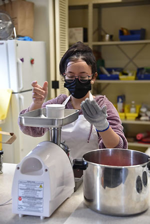 A student wearing an apron and gloves is standing behind a meat grinder in the kitchen.