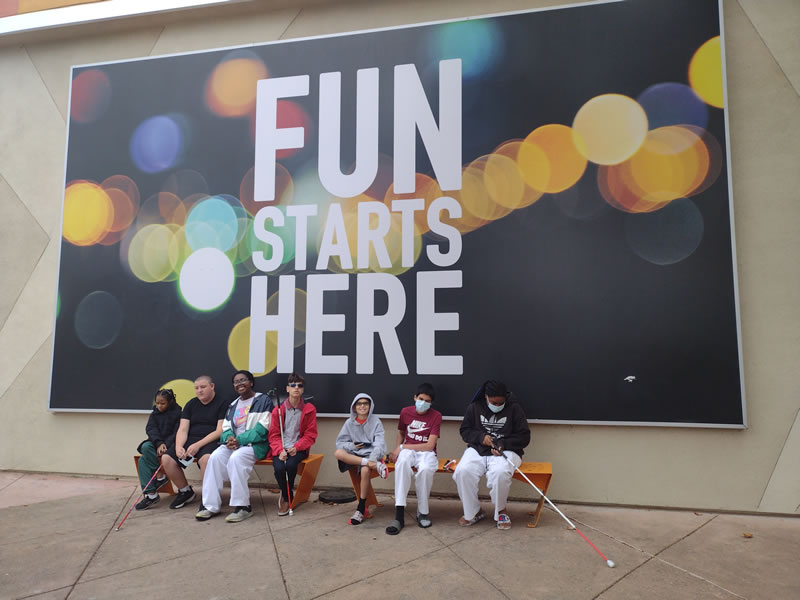 Seven California School for the Blind students sitting under a large banner with words, "fun starts here".
