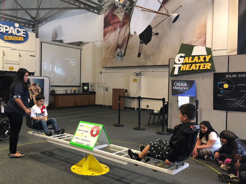 Two CSB students participating in an activity at a NASA exhibit with the help of a NASA guide. Both students are facing each other while sitting on a horizontal wide beam, which is elevated off of the ground. A few other students are pictured sitting around the exhibit, watching and listening to the two participants engaging in the activity.