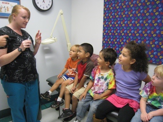 Several students sitting on a bench in a nurses office, as a nurse give instruction.