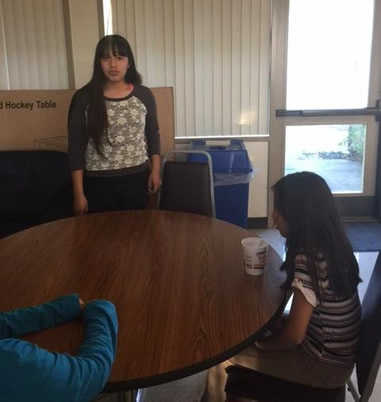 Student standing next to a table where other students are sitting and  listening to her.