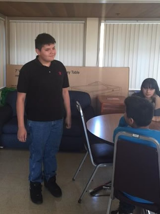 Student standing next to a table where other students are sitting and  listening to him.