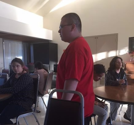 Student standing next to a chair, and other students sitting at tables in the background.