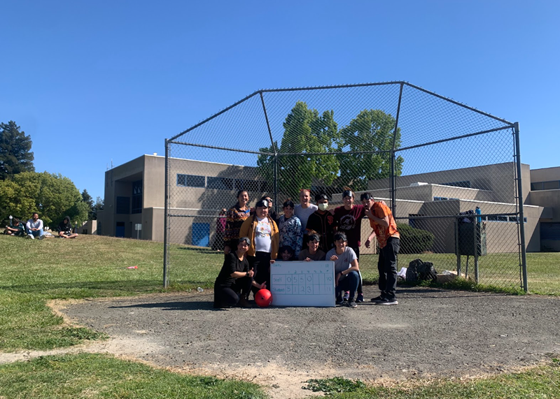 The CSB beep kickball team stands in a group in front of the backstop on the beep kickball field. The group is smiling behind a whiteboard that shows the points scored in each inning and the score of the game. 