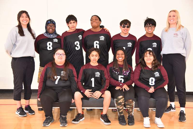 Four goalball players sit in the front row with five players behind them. Coach Taylor is standing on the left and coach Molly on the right. 