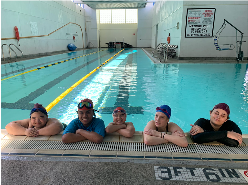 The 5 members of the CSB swim team are posing for a photo while in the pool. They have their arms resting on the pool deck and are all wearing a cap and goggles on their head. 