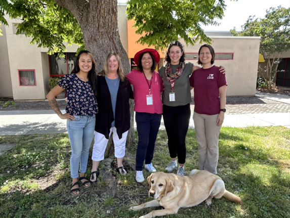 The 2023 O&M department poses arm in arm under the shade of a tree on the CSB campus. From left to right stands Laura Simpson, Kristi Barrella, Alicia Mendoza, Stacey Colley and Katie Marin. Laying down on the grass is Nanook, a CSB ambassador dog.